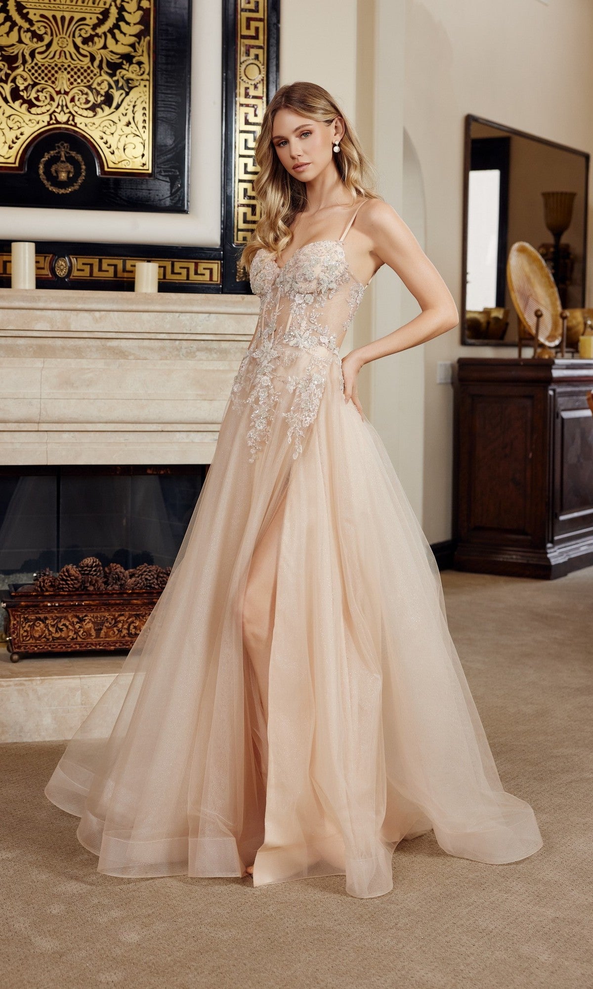 A woman stands in an elegant living room wearing the Nox Anabel G1787, a sheer blush floor-length prom dress with 3D floral appliques and a lace bodice.